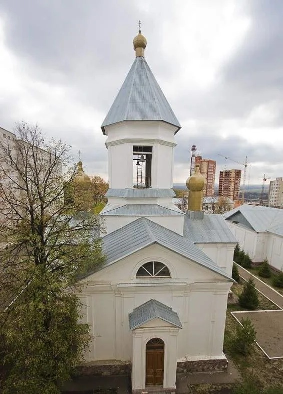 Белый храм с голубым куполом среди деревьев на фоне многоэтажных домов